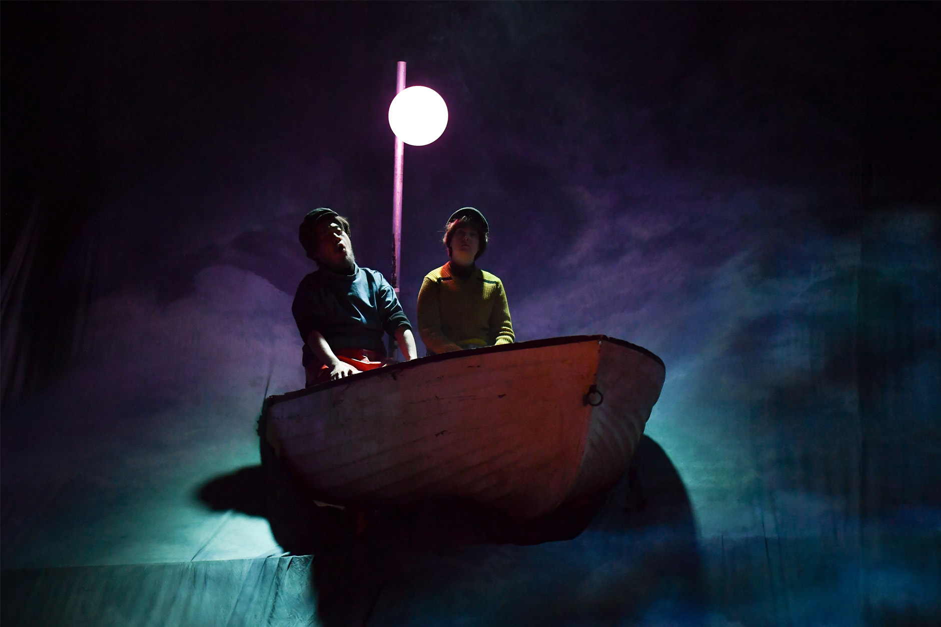 Two students, wearing fisherman costumes, sitting in a boat, singing on stage.